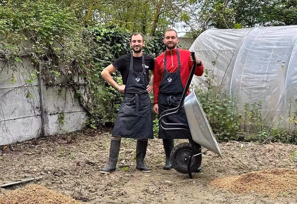 Deux brasseurs en tablier Léon transportent une brouette, devant un tas de drêche fraîche dans un jardin.